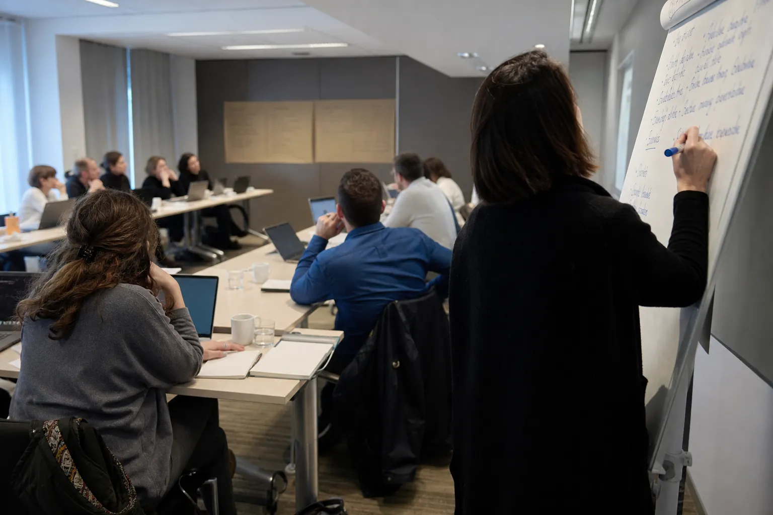 Salle de séminaire en séquence de travail, tables en U, participants de dos et paperboard couvert d’écriture