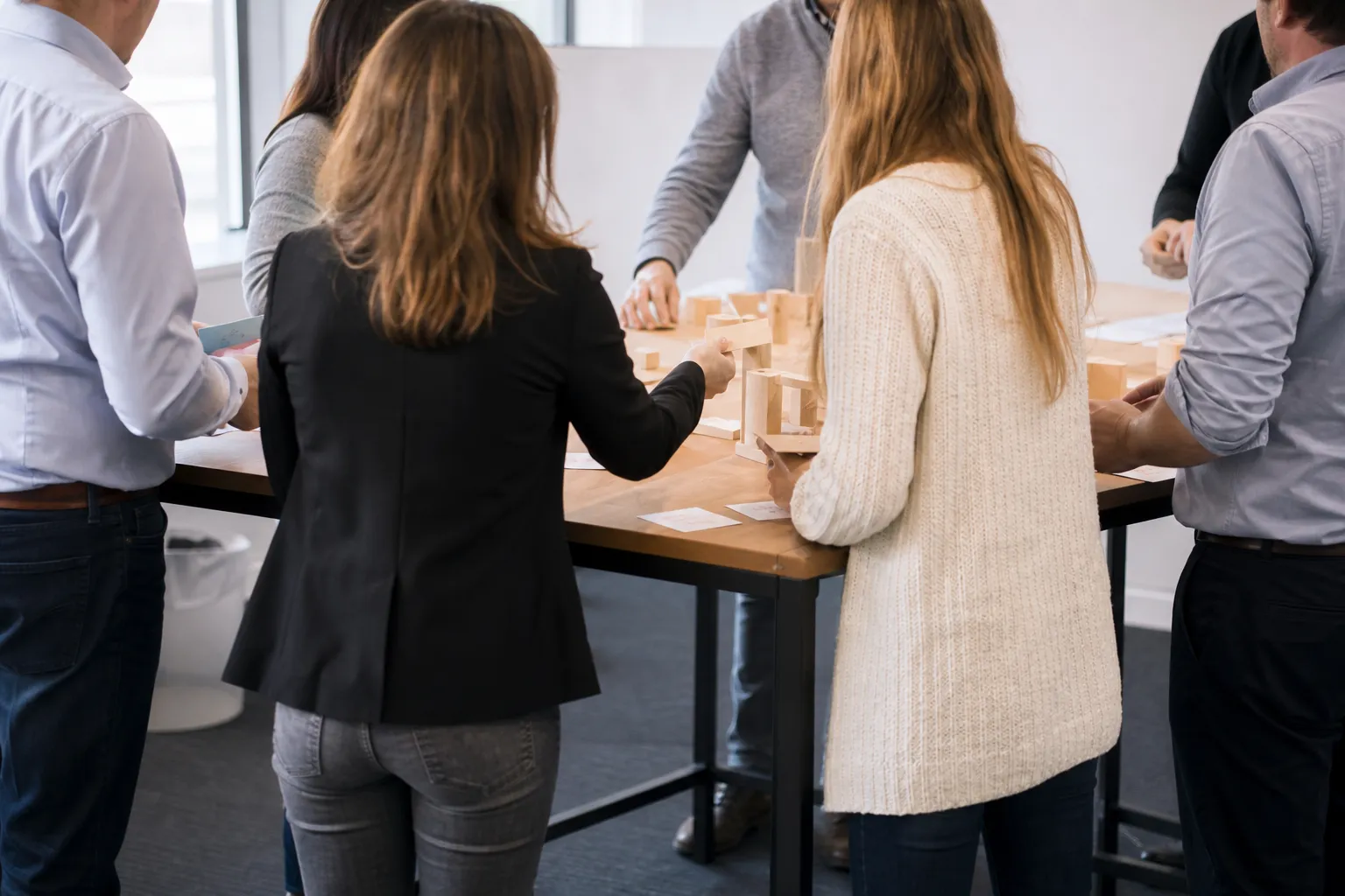 Atelier de cohésion autour d’une table haute, participants debout de dos manipulant des éléments de construction en bois